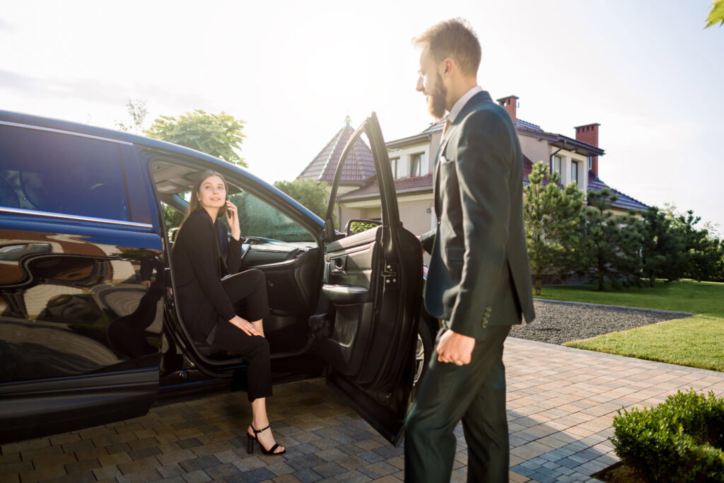 A woman in business attire sits in a car with the door open while a man in a suit approaches, outside a house on a sunny day.